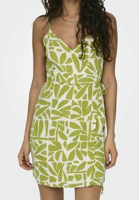 Robe portefeuille verte à motifs, décolletée en V, avec de fines bretelles, en tissu léger orné de formes de feuilles irrégulières sur un fond blanc.