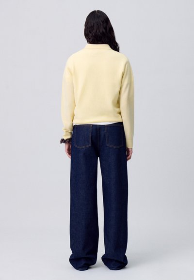 Suéter de punto amarillo con cuello, combinado con jeans de mezclilla oscuros de pierna ancha y alta cintura. La tela parece suave con detalles de costura visibles.