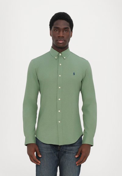 Chemise verte à manches longues en coton texturé, dotée d'un col classique et d'un petit logo bleu marine sur la poitrine. Portée avec un jean bleu.