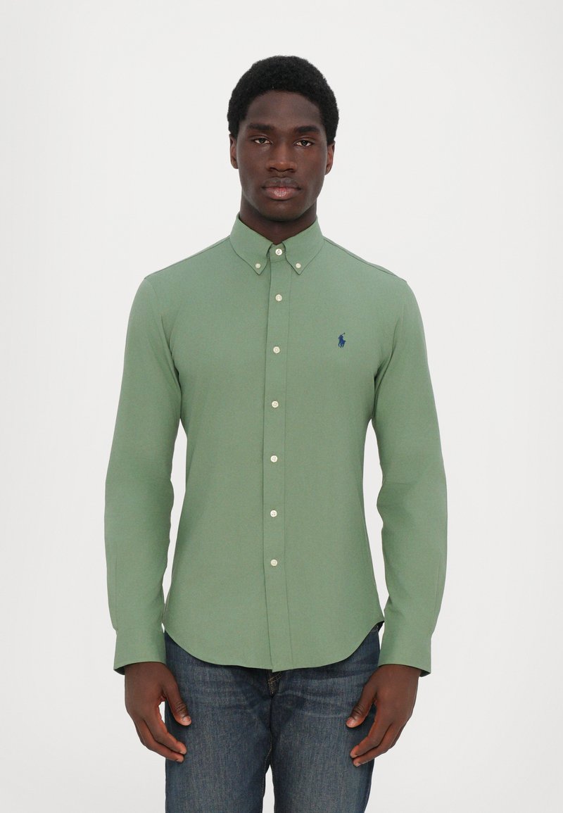 Chemise verte à manches longues en coton texturé, dotée d'un col classique et d'un petit logo bleu marine sur la poitrine. Portée avec un jean bleu.