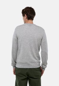 Homme portant un pull à manches longues gris clair et un pantalon vert foncé, se tenant de dos devant un fond blanc uni.