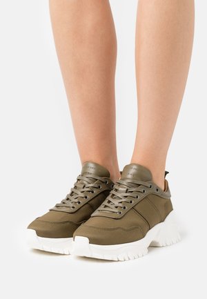 Sneakers verde oliva con upper in tela, punta tonda, suola in gomma bianca spessa e lacci piatti. Presenta un logo in rilievo sulla linguetta.
