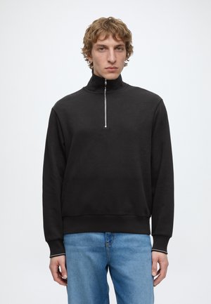 Jeune homme portant un sweat-shirt noir à fermeture éclair mi-longue et un jean bleu, debout devant un fond blanc uni.