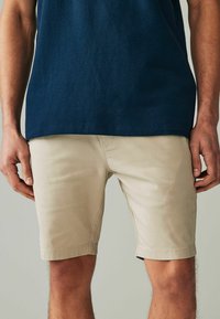 Beige pamut rövidnadrág slim fit szabással és feltekert szegéllyel. Sima textúra, elülső zsebek és rejtett gombos zipzárral, sötétkék inggel viselve.
