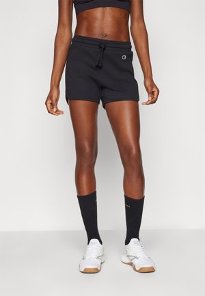 Pantalones cortos deportivos negros con cintura ajustable, con un parche de logo. Combinados con calcetines negros hasta la rodilla y zapatillas blancas.