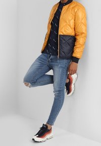 Gequilted oranje en marineblauwe bomberjack, marineblauw bedrukt T-shirt, versleten blauwe skinny jeans en chunky zwart/rode sneakers.