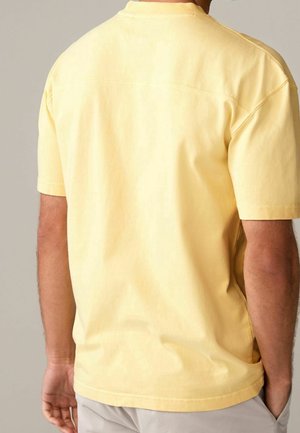 Homme portant un t-shirt à manches courtes jaune clair uni vu de dos, avec un pantalon beige clair, se tenant contre un fond neutre.