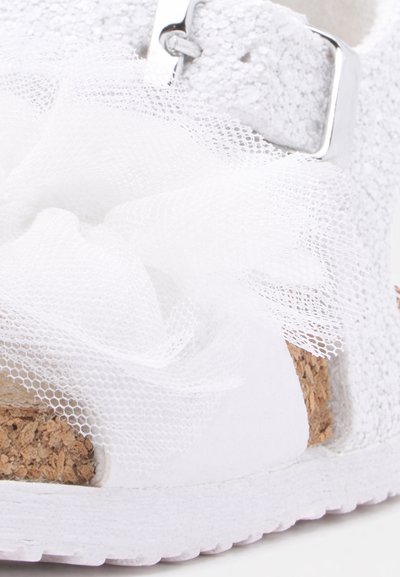 Κοντινό πλάνο ενός λευκού σανδαλιού με φελλό σόλα, ασημένιο λουράκι με αγκράφα και διακοσμητικό λευκό λουλούδι από δίχτυ ύφασμα στην κορυφή.