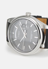 Montre Timex avec un boîtier en acier inoxydable, un cadran noir, des index en argent et un affichage jour-date. Elle est dotée d'un bracelet en cuir noir.