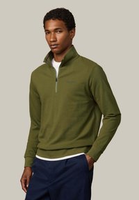 Suéter con cierre en verde oliva en un punto texturizado, con cuello alto y sutil marca 'HACKETT'. Lleva una camisa blanca debajo.