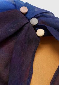 Blauwe en paarse doorschijnende stof draait samen, met drie decoratieve knopen in gedempte tinten. De achtergrond is van een gladde tankleurige materiaal.