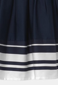 Marinblå kjol med veck och horisontella vita ränder vid fållen. Tillverkad av ett slätt tyg som framhäver en strukturerad form.