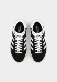 Svarta mockaskor med vita snören och tre vita sidostreck. Gummisula med texturerad tådel och Adidas-logotyp på tungan.