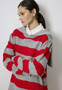 Pulover în dungi roșii și gri, cu glugă, textură moale de tricot, croială lejeră și mâneci lungi. Asortat cu o cămașă albă cu guler dedesubt.