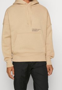 Hoodie en coton beige avec une poche kangourou, un capuchon avec cordon de serrage et un texte de logo brodé en marron foncé sur la poitrine gauche.