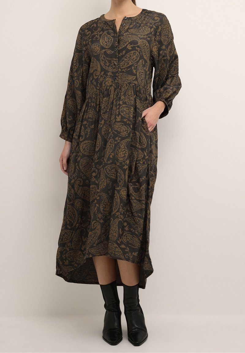 Femme portant une robe longue à manches longues de couleur sombre avec des motifs cachemire marron et des bottines noires, debout devant un fond neutre.