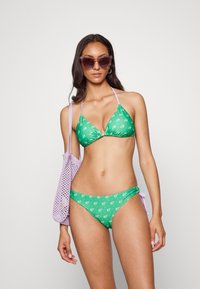 Bikini floral verde com um top triangular e alças ajustáveis; combinado com uma bolsa de malha roxa. Textura suave, cores vibrantes.