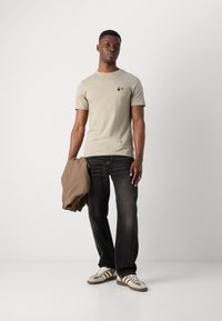 Gai bēšas krāsas īsām piedurknēm t-krekls ar nelielu izšūtu grafiku, saskaņots ar tumšiem džinsiem un gaiši brūnu jaku, papildināts ar daudzkrāsainiem sporta apaviem.