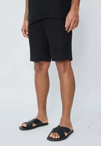 Shorts en lin noirs avec une coupe décontractée, associés à des sandales noires à lanières croisées. Design simple, tissu texturé et style minimaliste.
