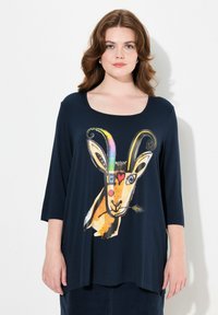 Kevyt, tummansininen paita, jossa on vuohigrafiikka värikkäillä yksityiskohdilla. Pyöreä kaula-aukko, kolmannen osan hihat ja rento leikkaus.