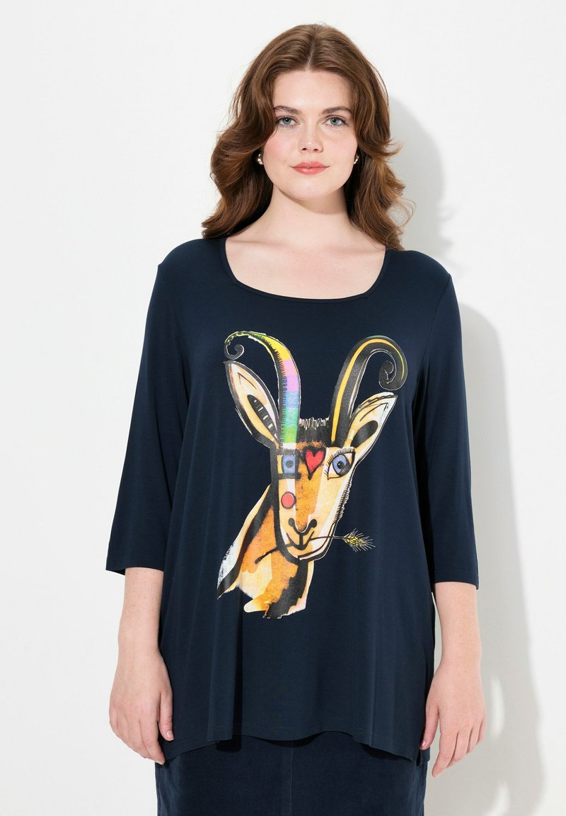 Kevyt, tummansininen paita, jossa on vuohigrafiikka värikkäillä yksityiskohdilla. Pyöreä kaula-aukko, kolmannen osan hihat ja rento leikkaus.