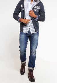 Felpa navy con zip e fodera grigia, testo bianco; camicia a quadri blu; jeans blu; stivali in pelle marrone con dettagli grigi.