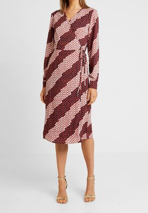 Frau trägt ein Wickelkleid mit langen Ärmeln in Midi-Länge, verziert mit diagonalen rötlich-braunen und weißen geometrischen Mustern, dazu beige Schuhe mit Knöchelriemen.