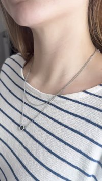 Colar em prata com várias correntes e um pequeno pendente circular com centro preto, usado sobre uma camisa às riscas brancas e azuis.