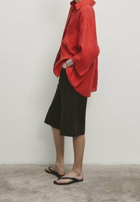 Chemise rouge surdimensionnée en tissu léger, associée à un short marron foncé jusqu'aux genoux et des tongs noires. Design simple, style décontracté.