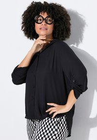 Schwarze Bluse mit Knopfleiste und Rolltab-Sleeves. Aus leichtem Stoff und mit lockerer Passform. Kombiniert mit schwarz-weißen karierten Hosen.