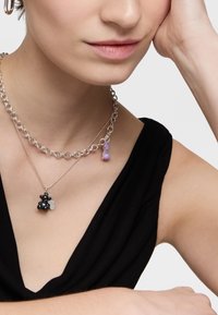 Donna che indossa due collane d'argento, una con un ciondolo a forma di orso nero e l'altra con una catena argentata e un charm viola, vestita con una maglietta nera.