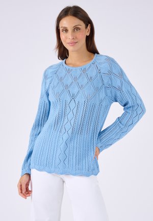 Vrouw die een lichtblauwe gebreide trui met lange mouwen draagt met diamant- en verticale patronen, gecombineerd met een witte broek, staand tegen een effen achtergrond.