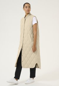 Long gilet beige matelassé avec un col montant, des fermetures à pression, des poches latérales et une finition mi-longue, porté sur une chemise blanche et un pantalon noir.