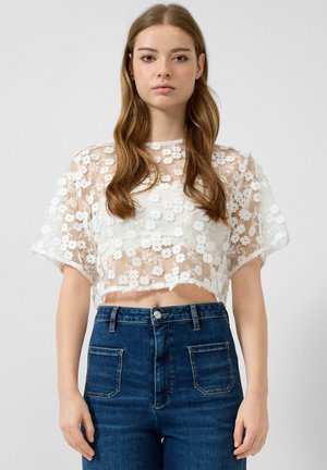 Młoda kobieta o długich włosach nosi przezroczysty biały kwiatowy koronkowy krótki top i niebieskie jeansy z wysokim stanem na jednolitym tle.