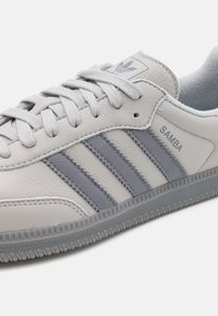 Bílé kožené tenisky se šedými akcenty, které mají tři šedé pruhy, strukturální gumovou podrážku a značkový štítek na jazyku.