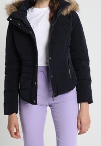 Tmavě námořnická péřová bunda s umělým kožíškem na kapuci, prošívanou texturou, zipovými kapsami a druky, kombinovaná s levandulovými kalhotami.