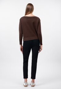 Woman standing facing away wearing a brown sweater, black slim-fit pants, and white sneakers on a plain white background.
