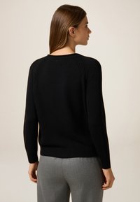 Schwarzer Strickpullover mit rundem Ausschnitt, gerippten Akzenten an den Schultern und einem figurbetonten Bündchen. Kombiniert mit grauen maßgeschneiderten Hosen.