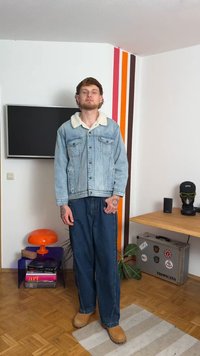 Denim jakna s krznim ovratnikom, svetlo modre barve, v kombinaciji z ohlapnimi temnimi modrimi kavbojkami in svetlimi čevlji. Ozadje vsebuje pisane stene s črtami.
