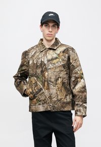 Ελαφρύ μπουφάν καμουφλάζ σε γήινους τόνους με υφή που μοιάζει με φλοιό δέντρου και φύλλα· διαθέτει γιακά και κλείσιμο με κουμπιά μπροστά.