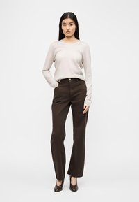 Femme aux longs cheveux noirs portant un haut beige à manches longues, un pantalon taille haute marron foncé et des chaussures à talons marron foncé, debout avec une main dans la poche.