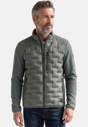 Mann mit grauem Haar und Bart trägt eine grüne gesteppte Jacke mit Reißverschluss und dunkle Jeans und steht vor einem schlichten Hintergrund.