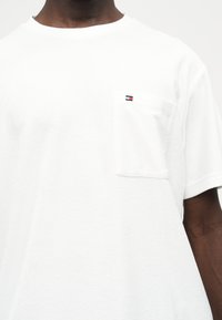 Άνδρας που φοράει απλό λευκό κοντομάνικο πουκάμισο με μικρό ορθογώνιο λογότυπο στην αριστερή τσέπη του στήθους, σε απλό φόντο.