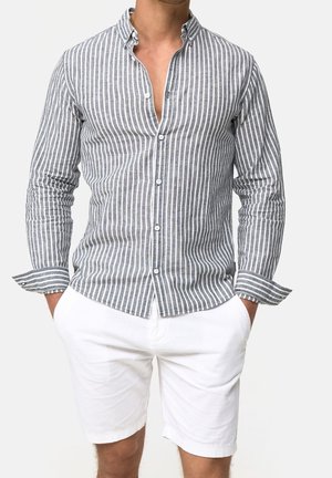 Hombre con camisa abotonada de rayas verticales grises y blancas, mangas arremangadas, y pantalones cortos blancos hasta la rodilla, con las manos en los bolsillos.