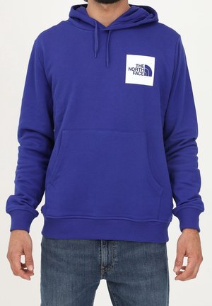 Mężczyzna ubrany w niebieską bluzę z kapturem i kieszenią z przodu oraz logo The North Face na klatce piersiowej, w połączeniu z niebieskimi jeansami.