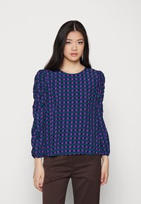 Top de mangas bufantes com padrão xadrez em roxo e verde sobre um fundo azul marinho. Apresenta um decote redondo e um tecido texturizado. Combinado com calças castanhas.