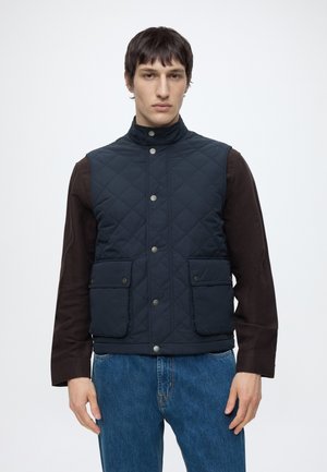 Jeune homme portant un gilet matelassé bleu marine sur une chemise sombre à manches longues et un jean bleu, debout devant un fond blanc uni.