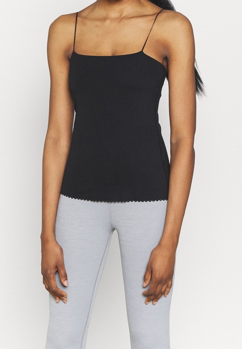 Femme portant un haut noir à fines bretelles et des leggings gris clair, debout devant un fond blanc uni.