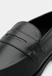 Crna kožna loaferica glatke završne obrade, s ukrasnim remenom i izrezima, prošivenim detaljima i zaobljenim oblikom vrha.
