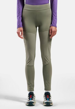 Olivegrüne legere Leggings mit hohem Bund, seitlichen Taschen und Knieflicken, kombiniert mit mehrfarbigen Sportschuhen.